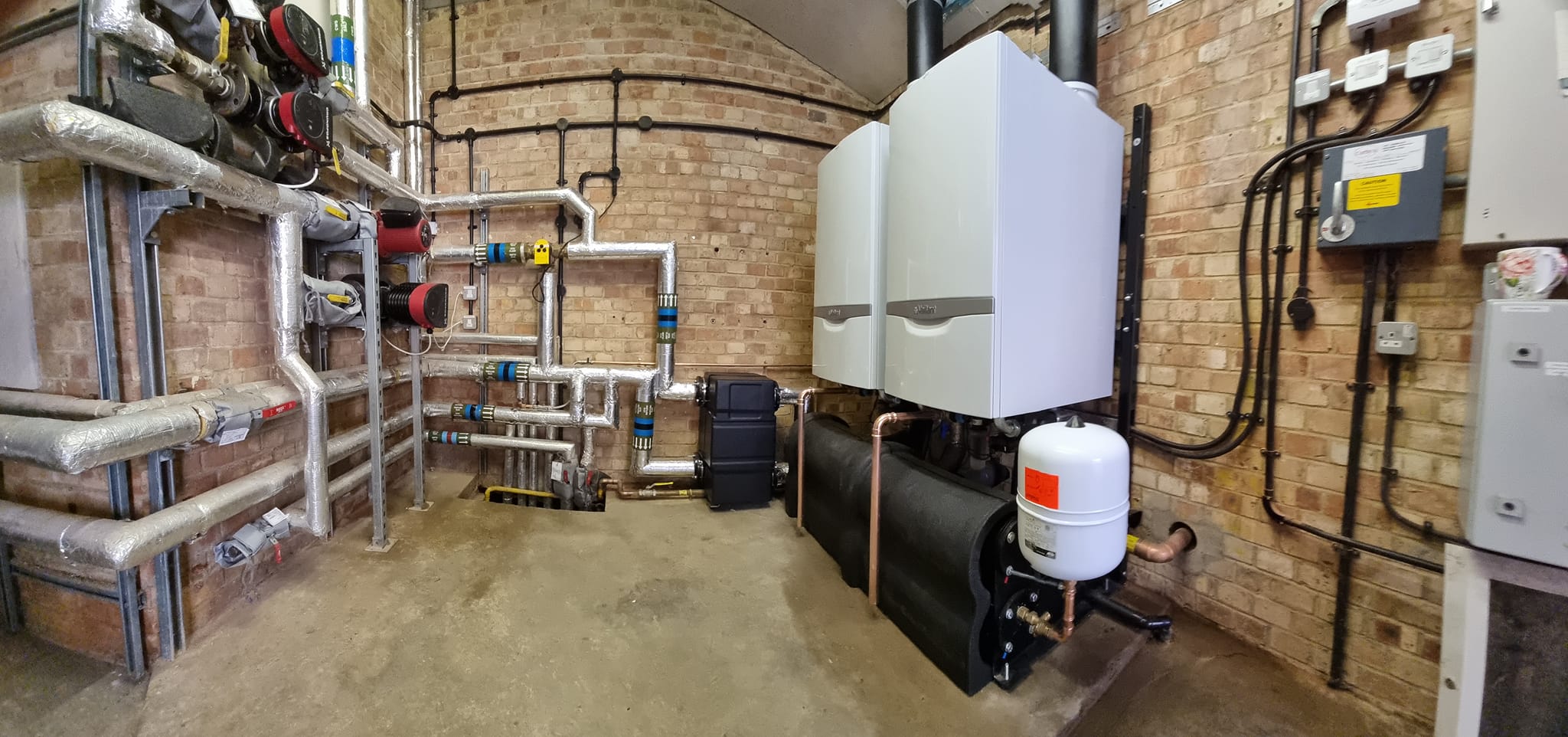 Commercial Boilers Care Home Commercial boiler system installed in a care home, featuring two large white boilers and intricate piping along the walls. The image shows a professional installation with insulated pipes and various control units.