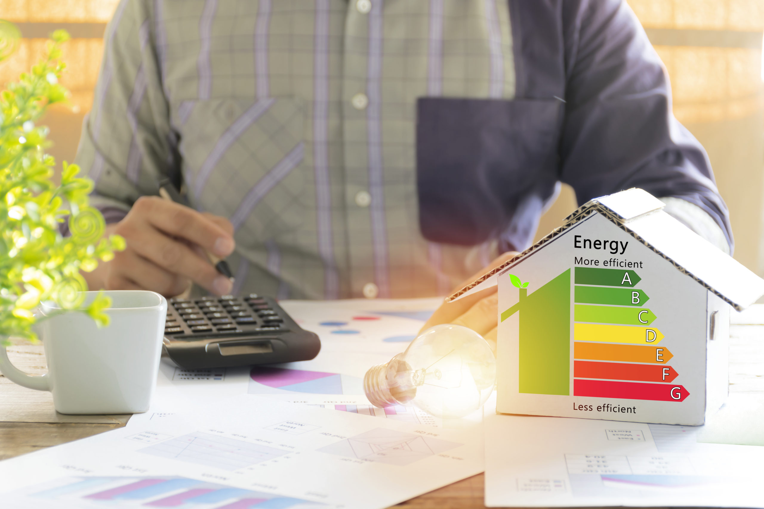 Man calculating energy savings with a model house displaying an energy efficiency rating, lightbulb, and financial charts on the table